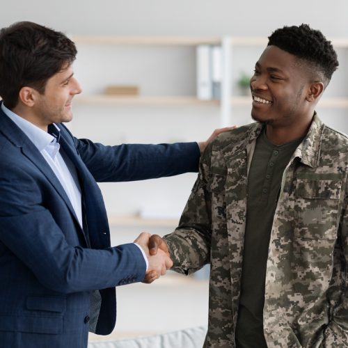 Cheerful guy in blue suit shaking happy young black military man hand, psychologist and soldier celebrating successful therapy, clinic interior. Military rehabilitation service concept